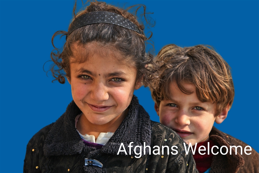 [ai] Two children smiling at the camera, with the girl in front wearing a black jacket and the boy behind with tousled hair. A blue background features the text "Afghans Welcome."